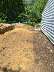 An excavation site next to a white building showing deep tire tracks in the orange-brown soil, a dug-out embankment, and lush green trees in the background